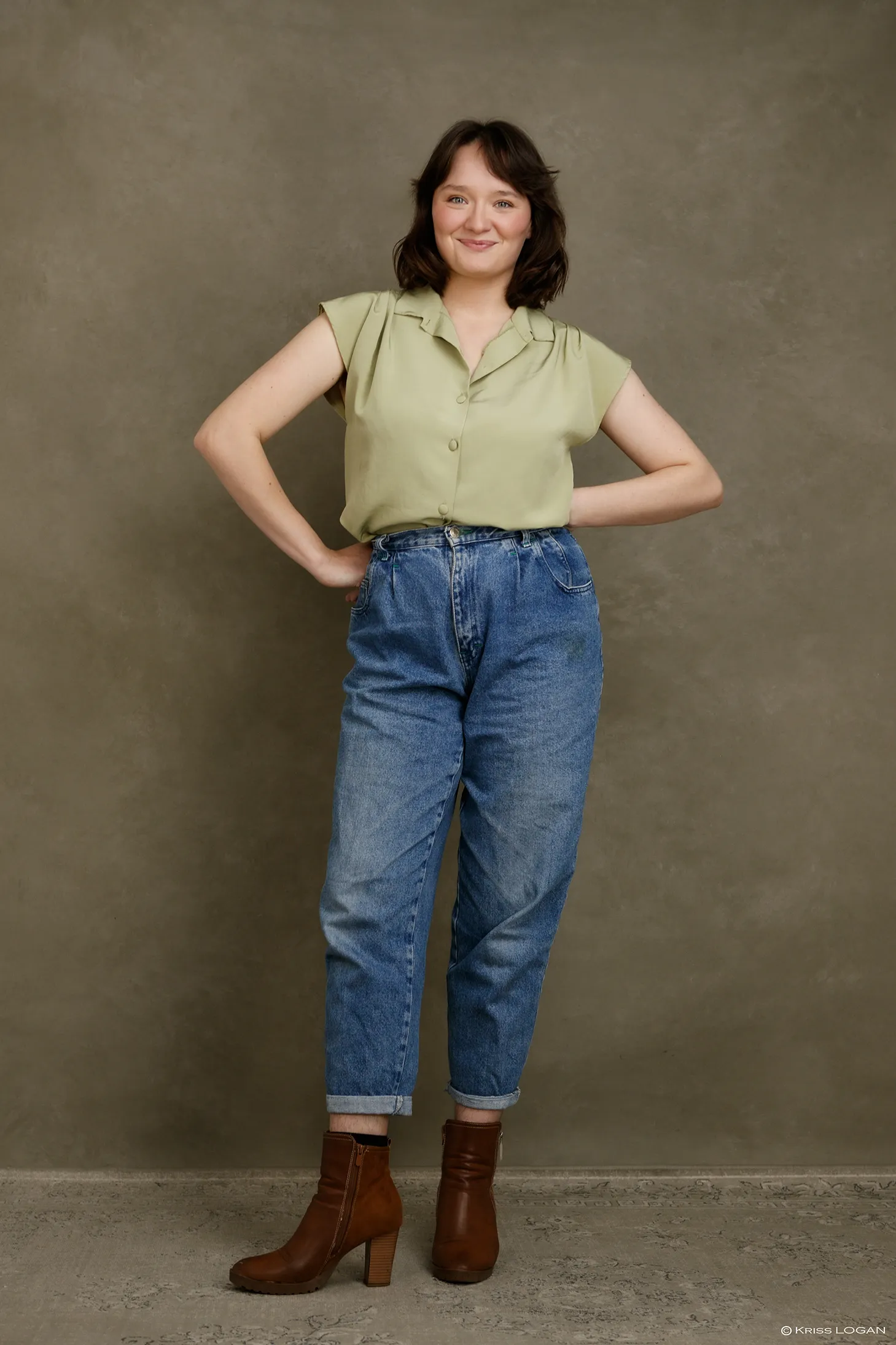 Plein-pied debout, mains aux hanches, chemise vert pâle et jean clair, bottines marron, fond sauge.