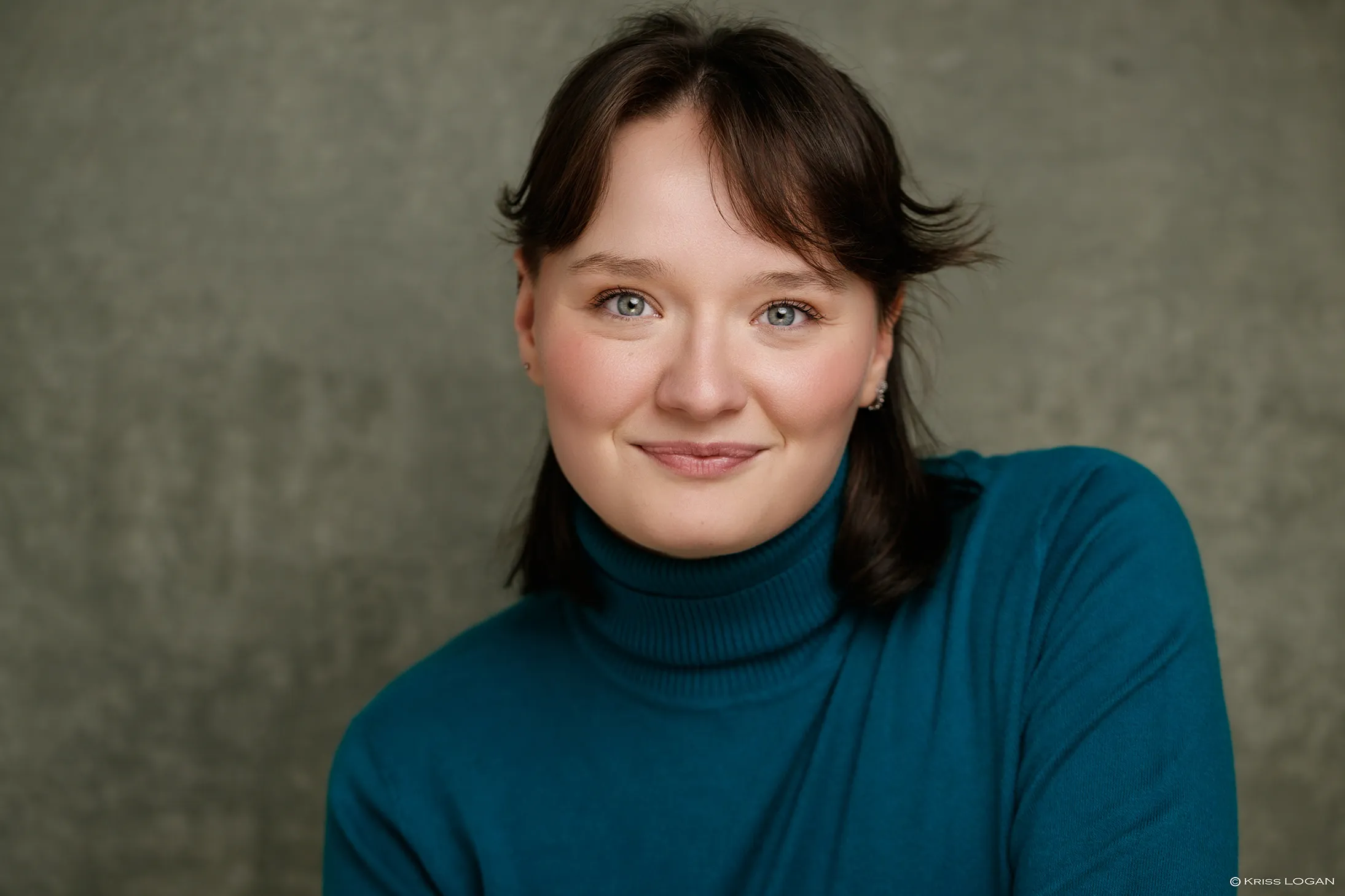 Portrait au sourire doux, pull col roulé bleu canard, fond gris-vert.