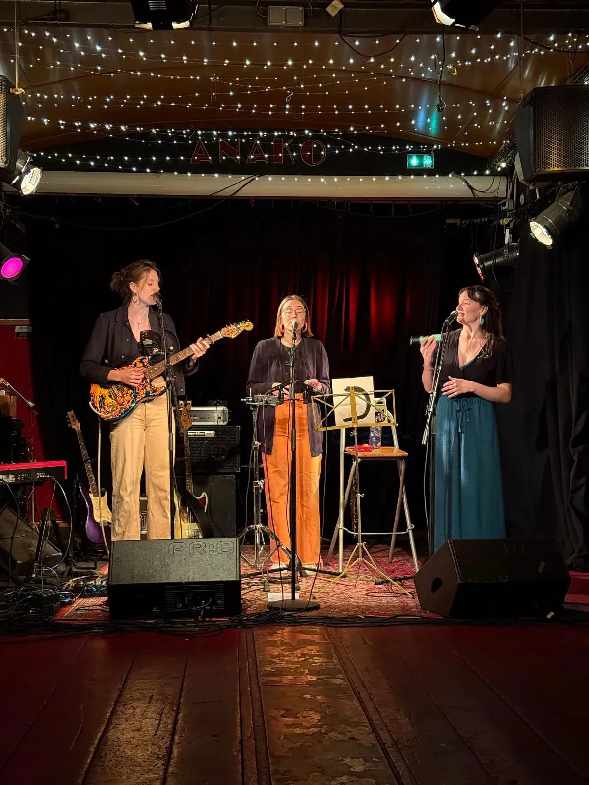 Trio on stage under a string of fairy lights, guitar and vocals.