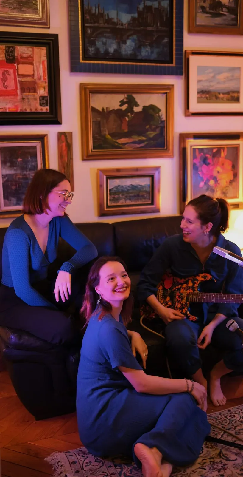Trio in a living room lined with old paintings, blue-tinted light.